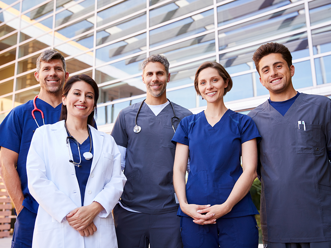 Group of smiling healthcare professionals and doctors standing outside modern medical facility in uniforms and lab coats