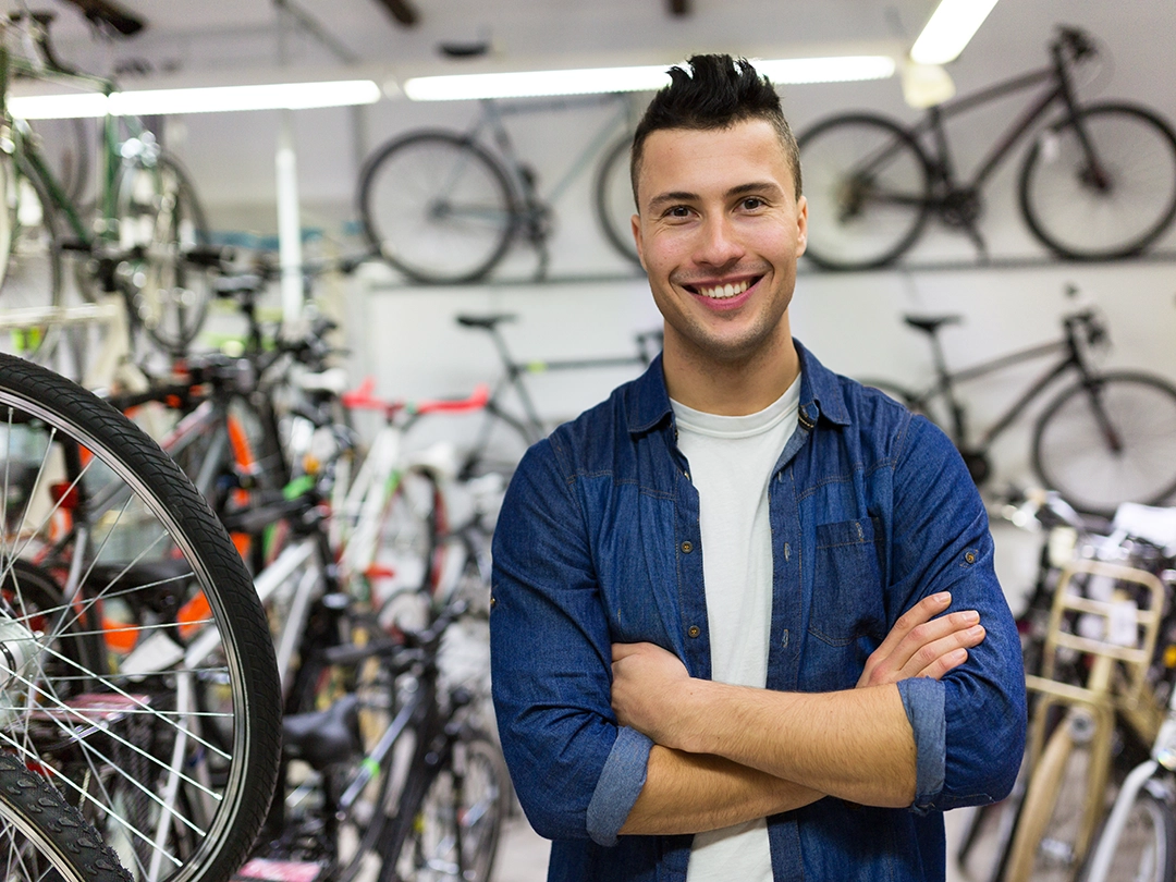 Smiling man standing with arms crossed inside a bicycle shop, surrounded by bikes, showcasing cycling retail and repair services.