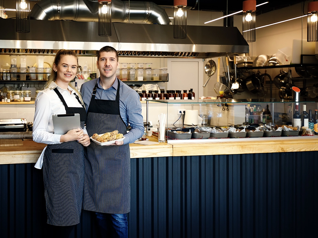 Smiling restaurant owners in aprons standing behind counter, holding food and tablet, inside modern kitchen.